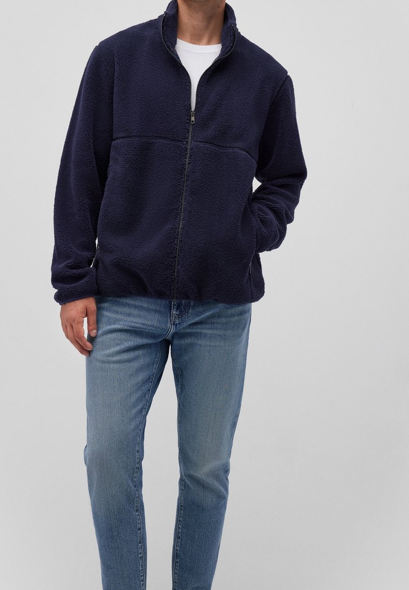 Giacca in pile blu navy con colletto alto e chiusura a mezza zip, con superficie testurizzata e due tasche laterali, abbinata a jeans azzurri.