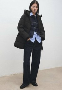 Czarna pikowana kurtka z kapturem, noszona na jeansowej kurtce, niebieskiej koszuli w paski, ciemnych dżinsach i czarnych butach. Prosty design z minimalnymi akcentami.