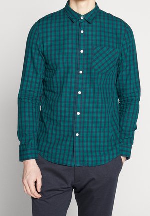 Homme portant une chemise à carreaux verte et bleue à manches longues avec une poche poitrine, associée à un pantalon foncé, sur un fond uni.