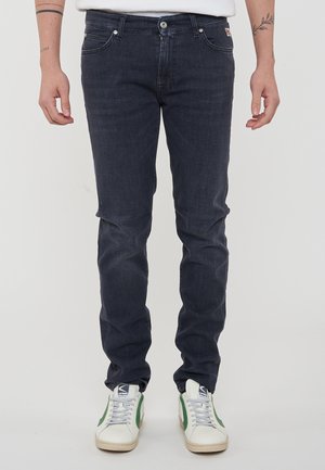 Jeans in denim blu scuro con una vestibilità affusolata, cinque tasche, zip e cuciture visibili. Indossati con sneakers bianche con accenti verdi.