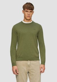 Olivgrüner Strickpullover mit Rundhalsausschnitt sowie gerippten Bündchen und Saum. Kleines Logo auf der Brust. Über einem weißen Hemd getragen, kombiniert mit hellen Hosen.