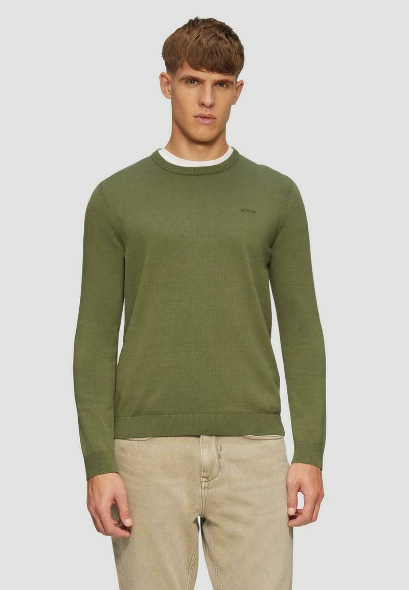 Olivgrüner Strickpullover mit Rundhalsausschnitt sowie gerippten Bündchen und Saum. Kleines Logo auf der Brust. Über einem weißen Hemd getragen, kombiniert mit hellen Hosen.