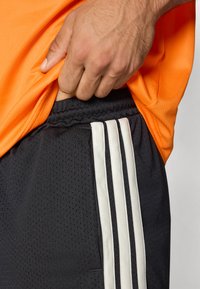 Black athletic shorts with white three-stripe design on the side, textured fabric, elastic waistband, worn with an orange shirt.
