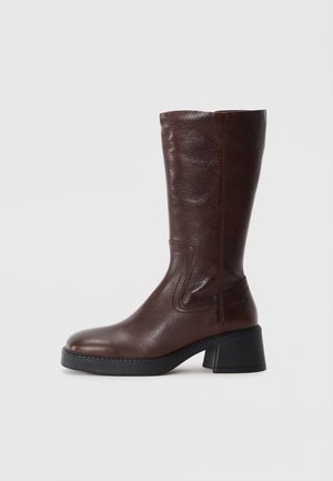 Bota de cuero marrón hasta la rodilla con un acabado texturizado, punta cuadrada y un tacón bajo cuadrado. Presenta costuras laterales y una parte superior ligeramente acampanada.