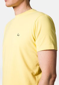 Żółta bawełniana koszulka z okrągłym dekoltem, krótkimi rękawami i małym czarnym logo na piersi. Miękka tekstura, swobodny krój.