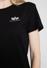 Schwarzes Baumwoll-T-Shirt mit kurzen Ärmeln, versehen mit einem weißen grafischen Logo und dem Text "ALPHA INDUSTRIES" im oberen linken Brustbereich.