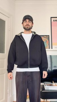 Muž v čiernej čiapke, čiernej bunde na zips cez bielu košeľu a tmavých nohaviciach, stojaci vo vnútri, v pozadí rámované umelecké diela a televízor.