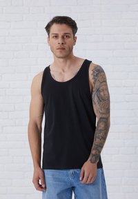 Zwarte tanktop van gladde stof, met brede armsgaten en een ronde hals, gecombineerd met lichte denim jeans en armtattoos.