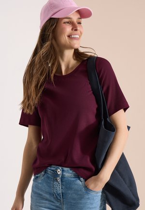 Femme souriante, portant une casquette rose, un t-shirt bordeaux à manches courtes, un jean bleu, et portant un sac cabas bleu marine sur l'épaule.