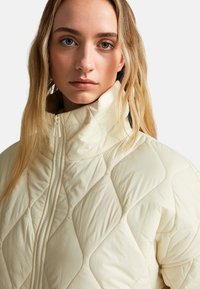 Světle béžová prošívaná bunda s vysokým límcem, zdobená diamantovými vzory, s předním zipem a hladkým, lesklým povrchem.