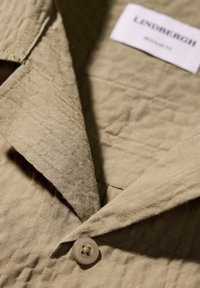 Camisa texturizada de color marrón claro con cuello clásico, que presenta un botón beige y una etiqueta que dice "Lindbergh."