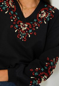 Schwarzer Sweatshirt mit buntem floralen Stickerei in Rot, Grün und Creme rund um den Halsausschnitt und die Bündchen, mit einem V-förmigen Design.