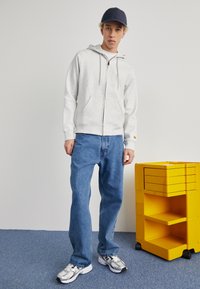 Grauer Reißverschluss-Hoodie mit Kordelzug, blaue Denim-Jeans und weiße Sneaker. Ein gelber Aufbewahrungswagen steht in der Nähe.