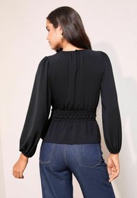Blusa nera con maniche a sbuffo e dettaglio in vita smock, realizzata in tessuto testurizzato. Abbinata a jeans in denim blu. Vista posteriore.