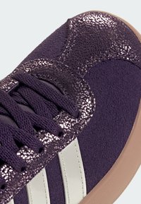 Sneaker viola con una parte superiore in suede viola testurizzato, accenti luccicanti, strisce bianche e una suola marrone chiaro. Design con lacci.