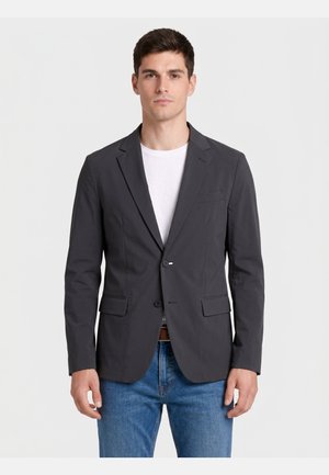 Młody mężczyzna stojący przodem, ubrany w ciemnoszary blezer na białej koszuli i niebieskich dżinsach na jasnym tle.