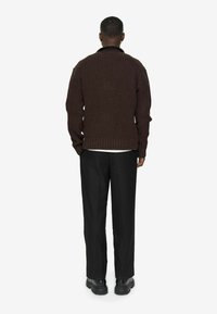 Barna kötött pulóver magas gallérral, bordás aljjal és hosszú ujjal, fekete nadrággal és fekete cipővel párosítva. Semleges háttér.