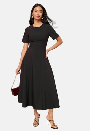 Black midi dress with short sleeves, fitted bodice, and flared skirt. Smooth fabric and subtle seams. Model holding a burgundy handbag.