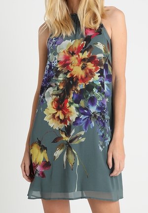 Robe grise sans manches avec un grand imprimé floral vibrant en jaune, rouge, violet et vert, portée par une personne aux cheveux blonds.