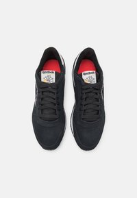 Ett par svarta Reebok Classic sneakers med svarta snören och röda innersulor, sedda ovanifrån på en vit bakgrund.