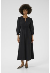 Black, long-sleeve dress with a keyhole neckline and gathered waist. Smooth fabric, ankle-length, paired with black and white striped heels.