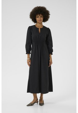 Black, long-sleeve dress with a keyhole neckline and gathered waist. Smooth fabric, ankle-length, paired with black and white striped heels.