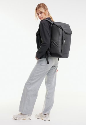 Kobieta w czarnej kurtce, jasnoszarych spodniach, białych trampkach, niosąca duży czarny plecak, spoglądająca przez ramię.