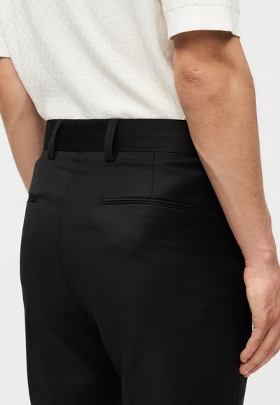 Κοντινό πλάνο ενός ατόμου που φοράει μαύρο παντελόνι με ραφή και λευκό υφασμάτινο πουκάμισο, εμφανίζοντας τις πίσω τσέπες και μέρος του δεξιού χεριού.