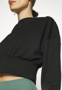 Sudadera recortada negra con un ajuste relajado, que presenta mangas largas y un dobladillo elástico. Textura de tela suave contra un fondo verde.