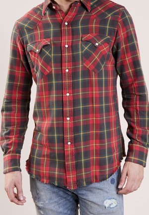 Uomo che indossa una camicia a quadri rossa e verde con due tasche sul petto e jeans blu consumati.