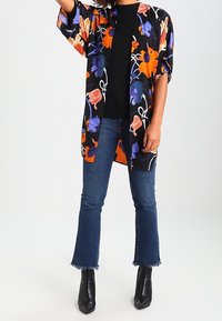 Floral kimono in het zwart met oranje, paarse en blauwe patronen. Losse pasvorm, mouwen tot halflang, gecombineerd met een zwart topje en uitlopende jeans. Zwarte enkellaarsjes.