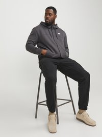 Grijze zip-up hoodie met capuchon, zwarte cargo broek en lichtbeige suède schoenen. Bevat een logo aan de voorkant en elastische manchetten op de broek.