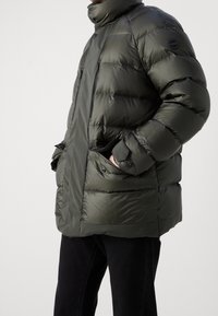 Chaqueta de plumas en verde oscuro, fabricada en tejido brillante. Presenta un alto cuello, bolsillos con cremallera y un diseño holgado con secciones acolchadas.