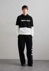Čierno-biela Moschino súprava s krátkou mikinou s nápisom a dlhými čiernymi nohavicami s bielym potlačou. Zmes bavlny, voľný strih.