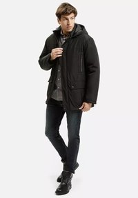 Czarna, ocieplona kurtka z kapturem, z frontowym zamkiem błyskawicznym, z kieszeniami na klapki, noszona na kraciastą koszulę, w zestawieniu z ciemnymi dżinsami i czarnymi butami.