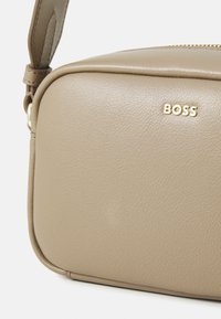 Kreminės odos per petį nešiojama piniginė su glotnia tekstūra, auksiniais aksesuarais ir prekės ženklo pavadinimu "BOSS" išspaustu ant priekio. Su užtrauktuku.