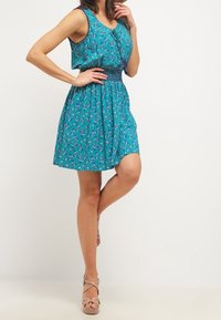 Robe florale bleu sarcelle avec une taille ajustée, un design sans manches et une jupe évasée. Tissu léger et respirant avec une texture froncée.