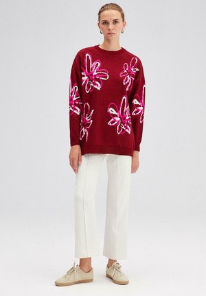 Bordeaux gebreide trui met roze en witte bloemenpatronen, gecombineerd met crèmekleurige broek met brede pijpen en lichtgekleurde sneakers.