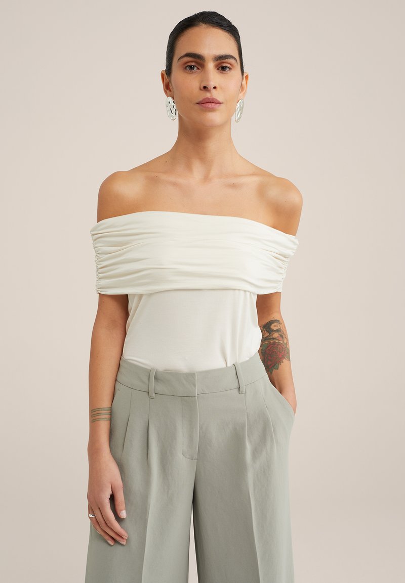 Top bianco scollato con dettagli di tessuto arricciato, abbinato a pantaloni grigi chiari a vita alta. Il modello ha un tatuaggio sul braccio.