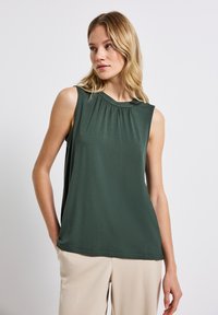 Canotta in tessuto verde scuro con scollo rotondo e dettaglio di pieghe frontali, abbinata a pantaloni leggeri beige. Texture liscia, design senza maniche.