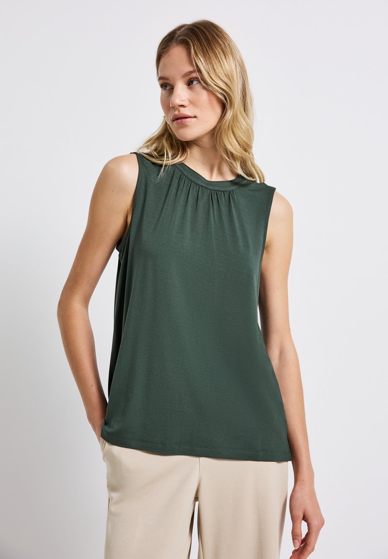 Canotta in tessuto verde scuro con scollo rotondo e dettaglio di pieghe frontali, abbinata a pantaloni leggeri beige. Texture liscia, design senza maniche.