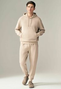 Hanorac bej și pantaloni de trening asortați din material moale, cu un buzunar frontal și manșete cu nervuri, purtați cu încălțăminte slip-on în nuanță taupe.