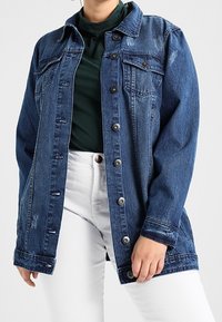 Jeansjacka, mörkblå, med en knapp-up front, två bröstfickor, lätt slitage och en avslappnad passform. Bärs över en grön topp.