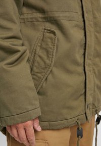 Olivno zelena izolirana jakna, ki vsebuje stranski žep, teksturirano tkanino, nastavljiv spodnji rob s vrvico in trdni zadrgo.