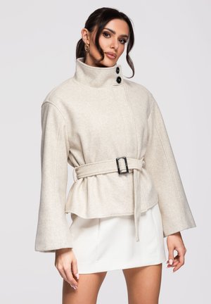 Frau trägt beige Wolljacke mit Gürtel, hohem Kragen und weiten Ärmeln, kombiniert mit weißem Minirock, posiert vor einfachem Hintergrund.