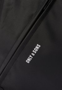 Tissu noir texturé avec une couture diagonale et le logo blanc "ONLY & SONS" imprimé près de la couture.
