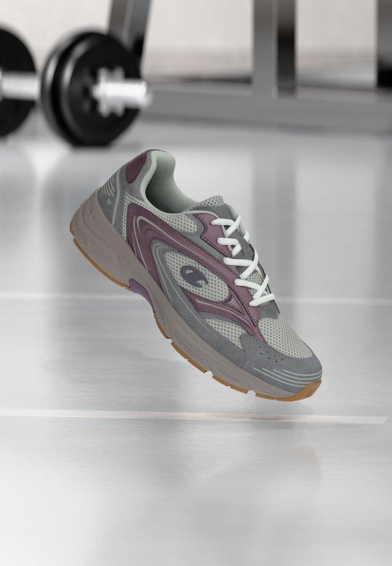 Γκρι και μωβ αθλητικό παπούτσι με λευκά κορδόνια να αιωρείται πάνω από το πάτωμα του γυμναστηρίου, με φλουταρισμένο αλτήρα και εξοπλισμό γυμναστηρίου στο φόντο.