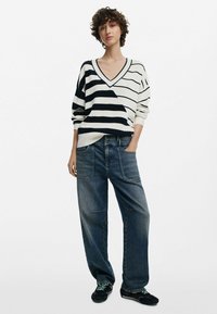 Maglione a righe, oversize, in crema e nero con scollatura a V; pantaloni in denim blu a vestibilità rilassata con grandi tasche; sneaker nere.