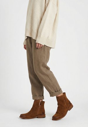 Personne portant un pull en maille beige, un pantalon marron retroussé et des bottines en daim marron, debout sur un fond blanc.