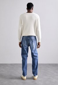 Vit ribbad stickad tröja, blå denimjeans med ljus tvätt och beiga skor, sedd bakifrån mot en enkel bakgrund.